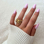 Una mano con uñas almendradas de color rosa claro y una manga de jersey de canalé blanco muestra varios anillos de oro apilados, entre ellos el elegante Anillo apilable brillante con diseños de textura y cuentas, que descansan sobre una superficie suave y neutra.
