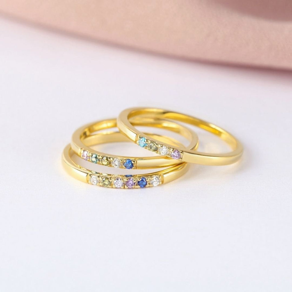 Tres anillos finos dorados con piedras de nacimiento personalizables se apilan sobre una superficie blanca con fondo rosa, ideales como anillo con piedras de nacimiento personalizado o un regalo especial para el Día de las Madres.