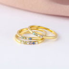 Tres anillos finos dorados con piedras de nacimiento personalizables se apilan sobre una superficie blanca con fondo rosa, ideales como anillo con piedras de nacimiento personalizado o un regalo especial para el Día de las Madres.