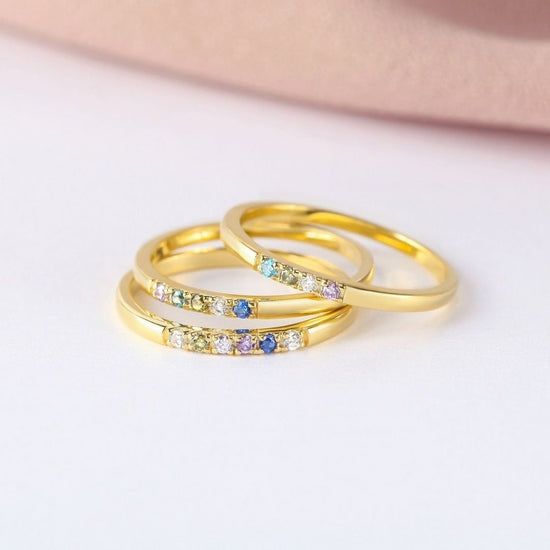 Tres anillos finos dorados con piedras de nacimiento personalizables se apilan sobre una superficie blanca con fondo rosa, ideales como anillo con piedras de nacimiento personalizado o un regalo especial para el Día de las Madres.