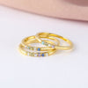 Tres anillos finos dorados con piedras de nacimiento personalizables se apilan sobre una superficie blanca con fondo rosa, ideales como anillo con piedras de nacimiento personalizado o un regalo especial para el Día de las Madres.
