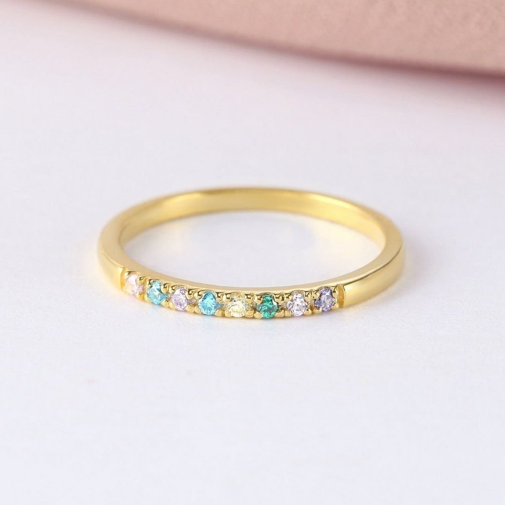 Un fino Anillo de oro con piedras de nacimiento personalizado que presenta una hilera de pequeñas piedras preciosas de colores engastadas a lo largo de la parte superior, expuestas sobre una superficie blanca con un suave fondo rosa.
