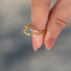 Una mano con uñas de color nude sujeta dos finos Anillos con piedras de nacimiento personalizados de oro, cada uno adornado con pequeñas piedras preciosas de colores, ideales como regalo personalizado o para el Día de las Madres. El fondo está suavemente difuminado en tonos fríos.
