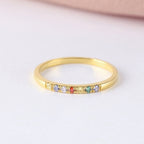 Un fino Anillo con piedras de nacimiento personalizado que presenta una hilera de pequeñas piedras preciosas de colores engastadas a lo largo de la mitad superior, expuestas sobre una superficie blanca con un suave fondo rosa.