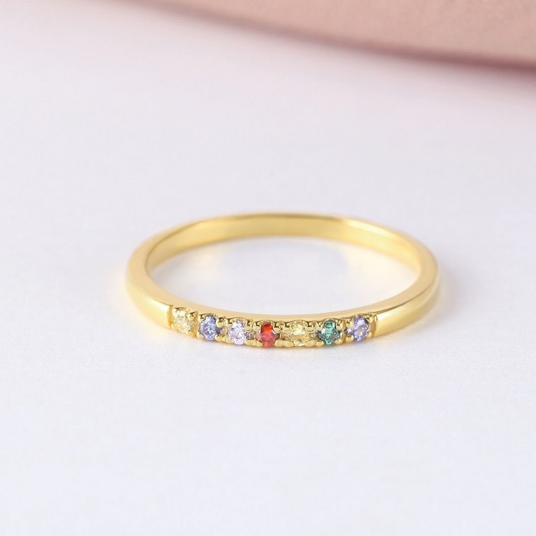 Un fino Anillo con piedras de nacimiento personalizado que presenta una hilera de pequeñas piedras preciosas de colores engastadas a lo largo de la mitad superior, expuestas sobre una superficie blanca con un suave fondo rosa.