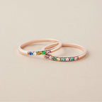 Dos elegantes Anillos con piedras de nacimiento personalizados en oro rosa, cada uno con pequeñas gemas de colores en una sola hilera, exhibidos sobre una superficie beige clara.