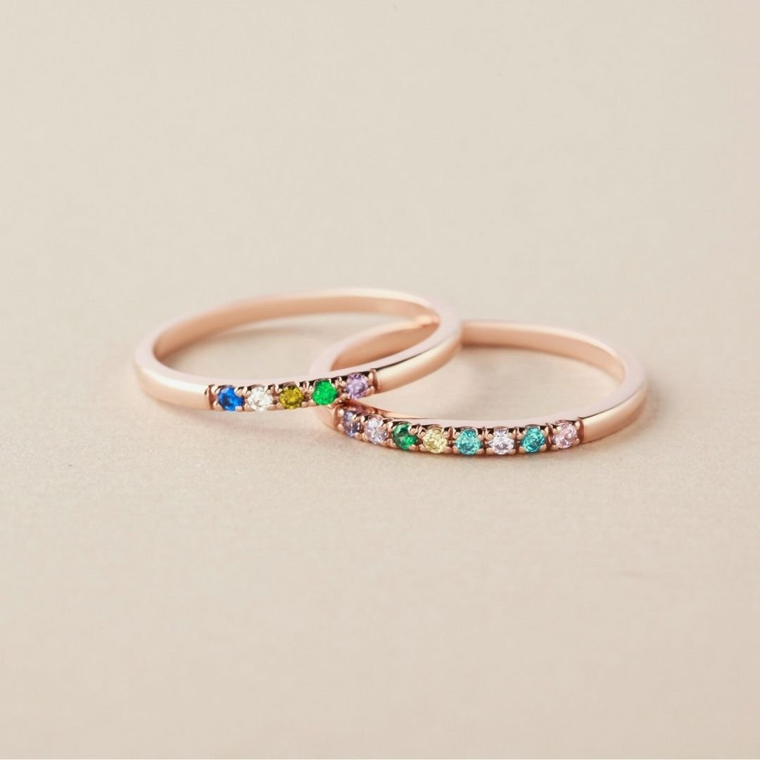 Dos elegantes Anillos con piedras de nacimiento personalizados en oro rosa, cada uno con pequeñas gemas de colores en una sola hilera, exhibidos sobre una superficie beige clara.