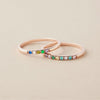 Dos elegantes Anillos con piedras de nacimiento personalizados en oro rosa, cada uno con pequeñas gemas de colores en una sola hilera, exhibidos sobre una superficie beige clara.