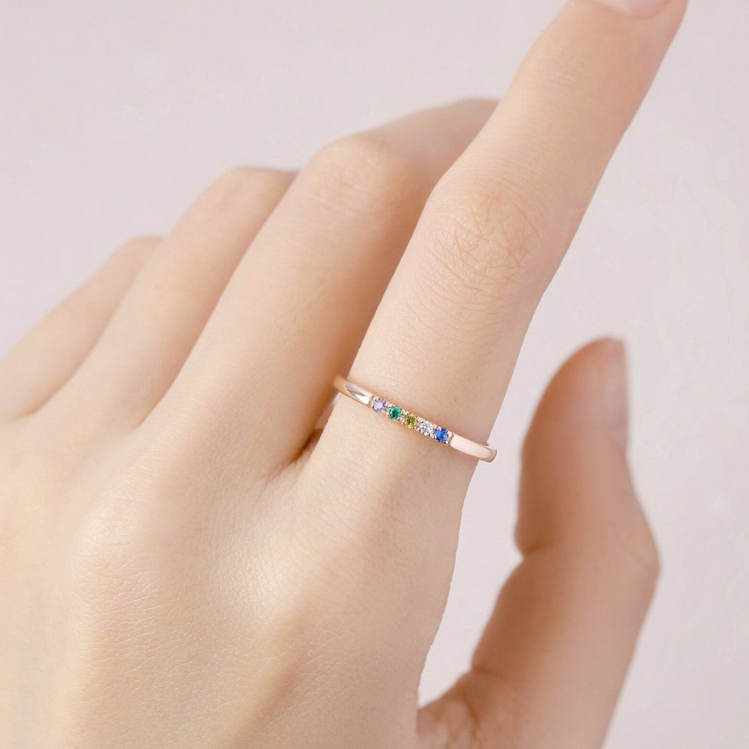 Una mano de piel clara luce en el dedo anular el Anillo con piedras de nacimiento personalizado, con pequeñas gemas de colores. El fondo suavemente difuminado resalta el delicado diseño personalizado del anillo.