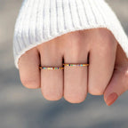 Una mano vestida con un jersey blanco de canalé muestra dos anillos de oro con pequeñas piedras preciosas de colores, entre ellas el Anillo con piedras de nacimiento personalizado, que lleva en los dedos corazón y anular. Las uñas están pintadas de color nude.