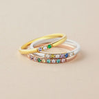 Tres delicados anillos -oro, plata y oro rosa- adornados con vibrantes piedras preciosas inspiradas en el Anillo con piedras de nacimiento personalizado, se apilan con elegancia sobre una superficie beige.