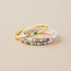 Tres delicados anillos -oro, plata y oro rosa- adornados con vibrantes piedras preciosas inspiradas en el Anillo con piedras de nacimiento personalizado, se apilan con elegancia sobre una superficie beige.