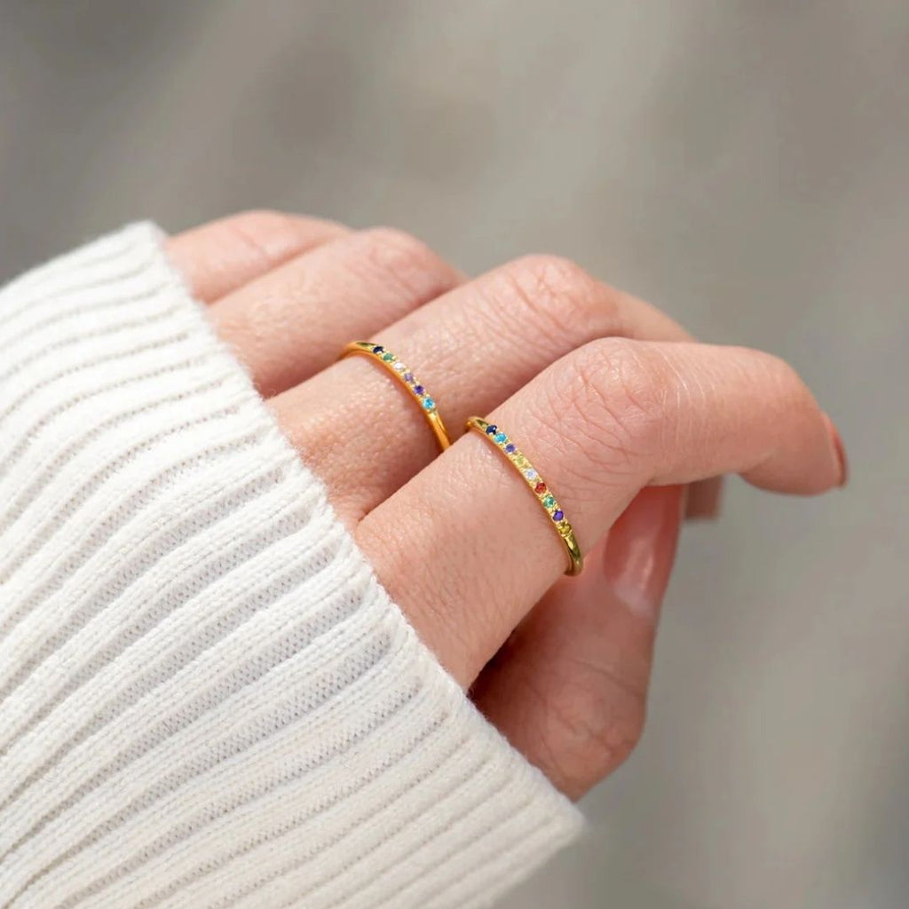 Primer plano de una mano que lleva dos finos anillos de oro con piedras de nacimiento personalizadas, cada uno engastado con pequeñas gemas de colores. La persona lleva un jersey blanco de canalé, mientras que el fondo suavemente difuminado resalta el encanto de los anillos.