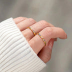 Primer plano de una mano que lleva dos finos anillos de oro con piedras de nacimiento personalizadas, cada uno engastado con pequeñas gemas de colores. La persona lleva un jersey blanco de canalé, mientras que el fondo suavemente difuminado resalta el encanto de los anillos.