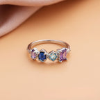 Anillo con piedras natales en plata, personalizado con cuatro piedras en tonos lila, azul oscuro, aqua y lavanda. Presentado sobre superficie durazno y fondo de tela rosa suave. Ideal como regalo para el Día de las Madres.
