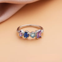 Anillo con piedras natales en plata, personalizado con cuatro piedras en tonos lila, azul oscuro, aqua y lavanda. Presentado sobre superficie durazno y fondo de tela rosa suave. Ideal como regalo para el Día de las Madres.