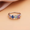 Anillo con piedras natales en plata, personalizado con cuatro piedras en tonos lila, azul oscuro, aqua y lavanda. Presentado sobre superficie durazno y fondo de tela rosa suave. Ideal como regalo para el Día de las Madres.