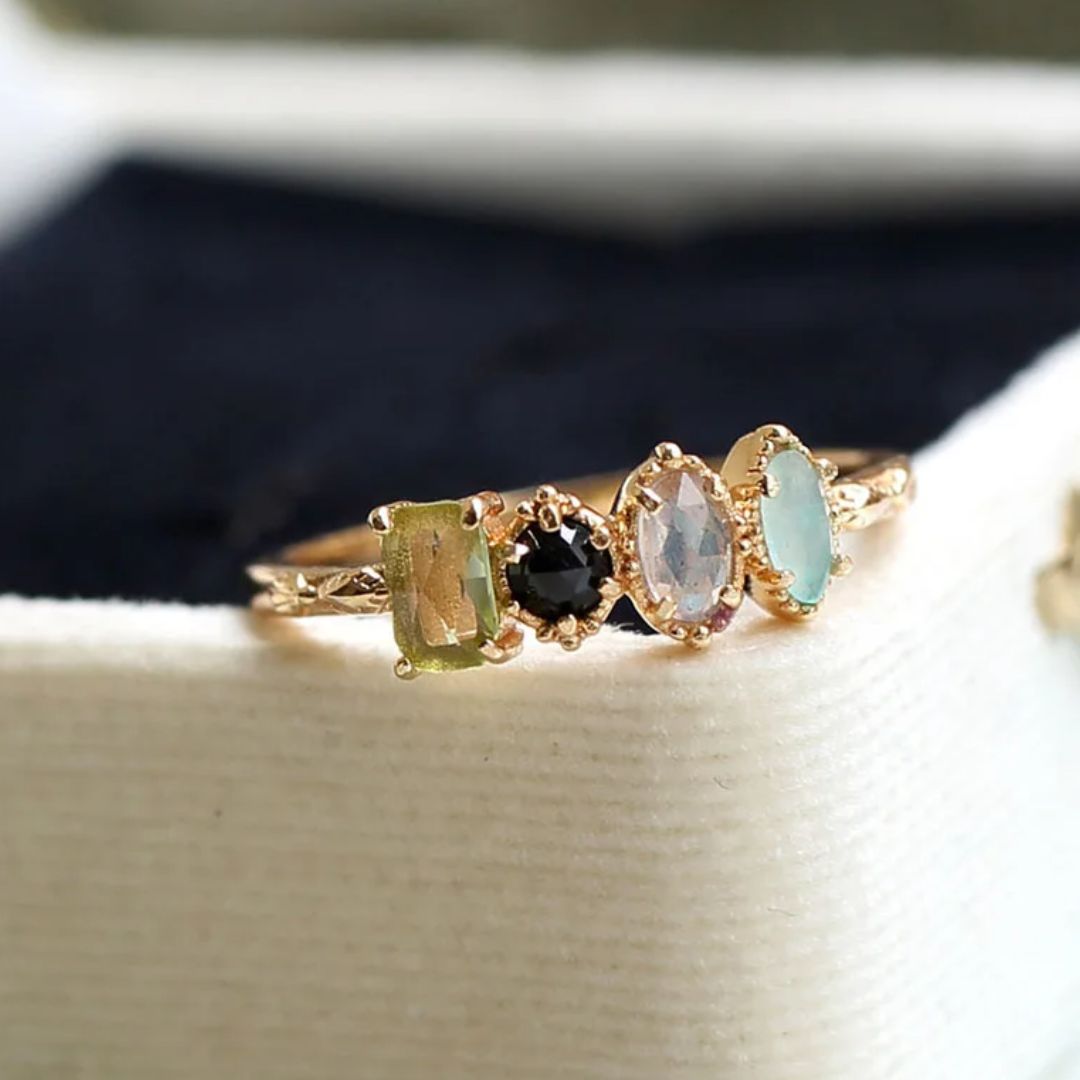 El Anillo con piedras natales de oro luce cuatro gemas únicas en distintos colores y formas, presentadas sobre tela crema y fondo oscuro; un regalo ideal para el Día de las Madres.