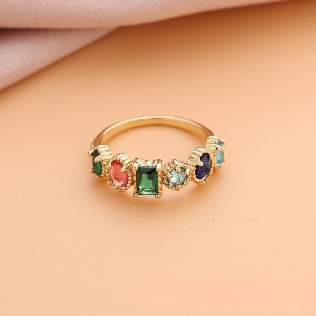 El Anillo con piedras natales de oro luce cinco gemas de colores-verde, rojo, dos celestes y azul oscuro-alineadas sobre fondo durazno y tela beige. Un regalo perfecto para el Día de las Madres.