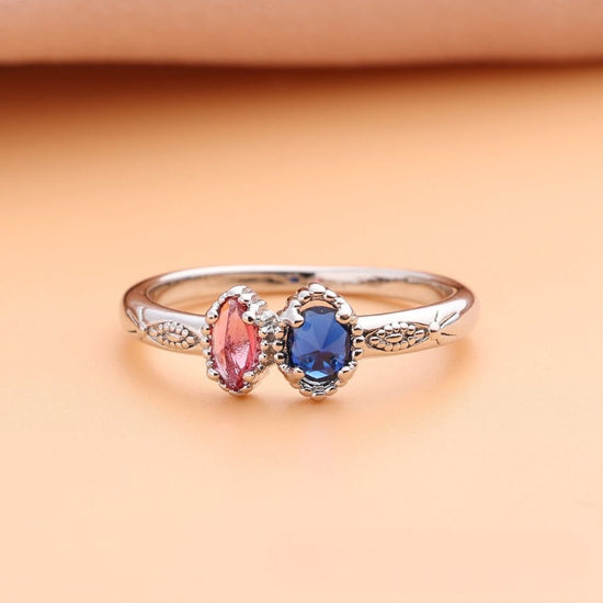 Un delicado anillo con piedras natales color plateado, con dos gemas ovaladas (rosa y azul) y detalles intrincados-ideal como anillo piedras de nacimiento o regalo Día de las Madres. Se muestra en una suave superficie melocotón.