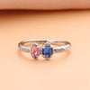 Un delicado anillo con piedras natales color plateado, con dos gemas ovaladas (rosa y azul) y detalles intrincados-ideal como anillo piedras de nacimiento o regalo Día de las Madres. Se muestra en una suave superficie melocotón.