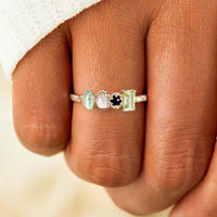 Un primer plano de una mano luciendo el Anillo con piedras natales, personalizado en plata con cuatro piedras de diferentes formas y colores (verde claro, blanco y negro); un regalo perfecto para el Día de las Madres.