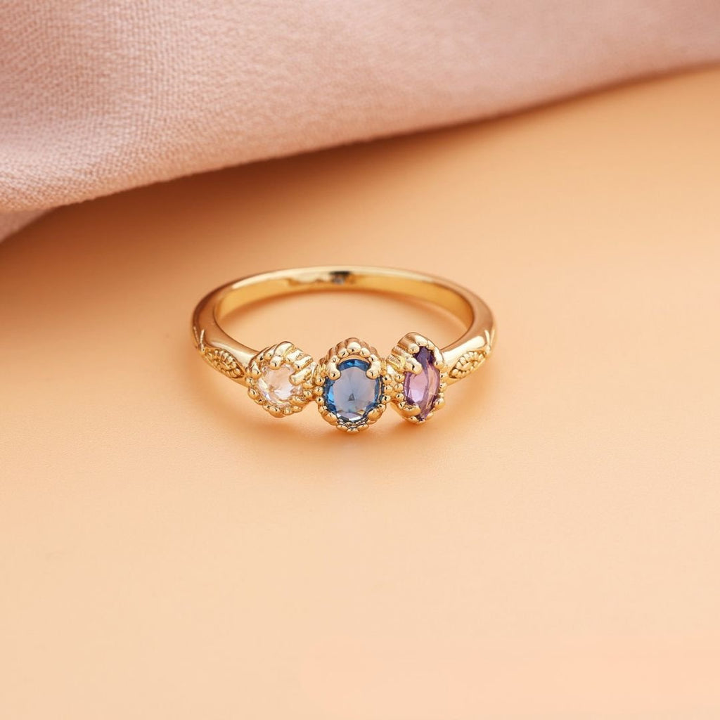 Anillo con piedras natales de oro con tres gemas ovaladas rosa, azul y morado, alineadas sobre una superficie durazno y tela rosada suave; el regalo ideal para el Día de las Madres.