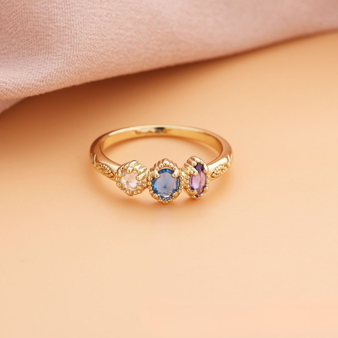 Anillo con piedras natales de oro con tres gemas ovaladas rosa, azul y morado, alineadas sobre una superficie durazno y tela rosada suave; el regalo ideal para el Día de las Madres.