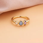 Anillo con piedras natales de oro con tres gemas ovaladas rosa, azul y morado, alineadas sobre una superficie durazno y tela rosada suave; el regalo ideal para el Día de las Madres.