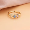 Anillo con piedras natales de oro con tres gemas ovaladas rosa, azul y morado, alineadas sobre una superficie durazno y tela rosada suave; el regalo ideal para el Día de las Madres.