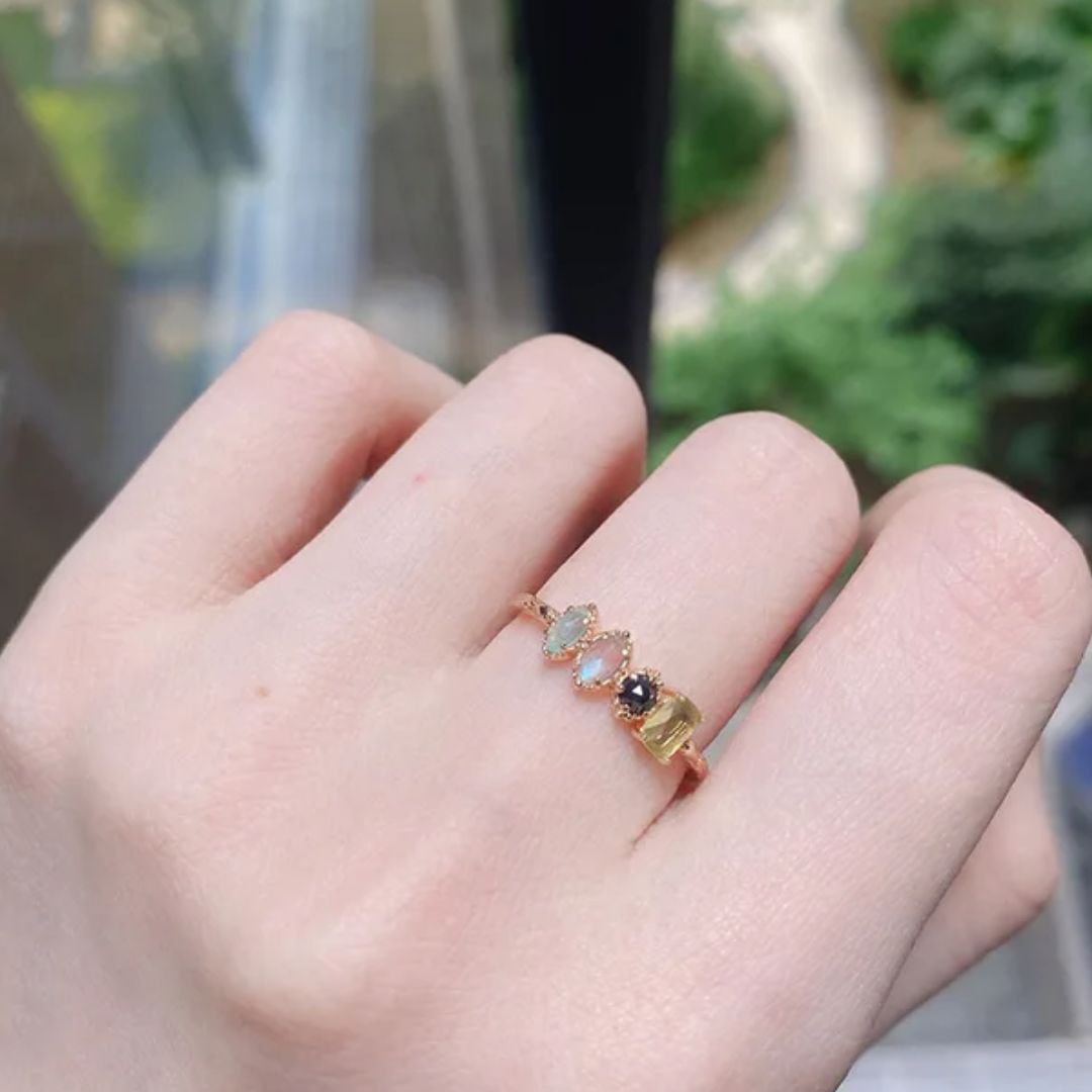 Una mano luce un delicado Anillo con piedras natales-engarzado con cinco piedras preciosas de colores-brillando sobre un verdor difuminado. Ideal como un anillo personalizado o un regalo Día de las Madres.