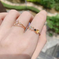 Primer plano de una mano que lleva dos delicados anillos de oro, entre ellos el Anillo con piedras natales, con pequeñas gemas de colores. La vegetación difuminada y un camino al fondo sugieren un entorno al aire libre.