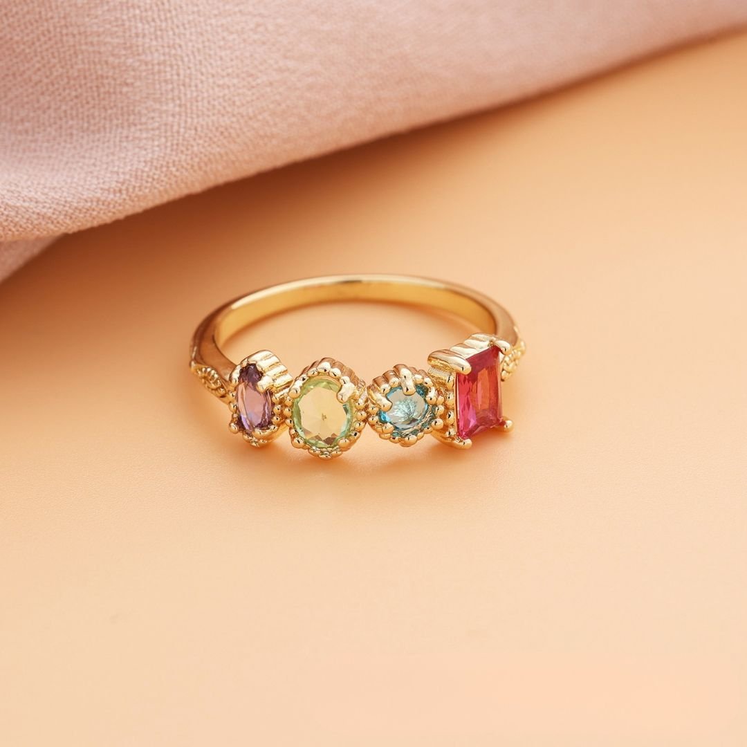 El Anillo con piedras natales en oro luce cuatro gemas de nacimiento-púrpura, amarillo, azul y rojo-en engastes ornamentados, sobre fondo durazno y tela rosa; ideal como regalo para el Día de las Madres.