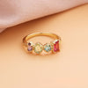 El Anillo con piedras natales en oro luce cuatro gemas de nacimiento-púrpura, amarillo, azul y rojo-en engastes ornamentados, sobre fondo durazno y tela rosa; ideal como regalo para el Día de las Madres.