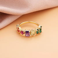 Un Anillo con piedras natales de oro con seis piedras preciosas rectangulares en morado, rojo, amarillo y verde descansa sobre una superficie melocotón con suave tela rosa-ideal como regalo para el Día de la Madre.