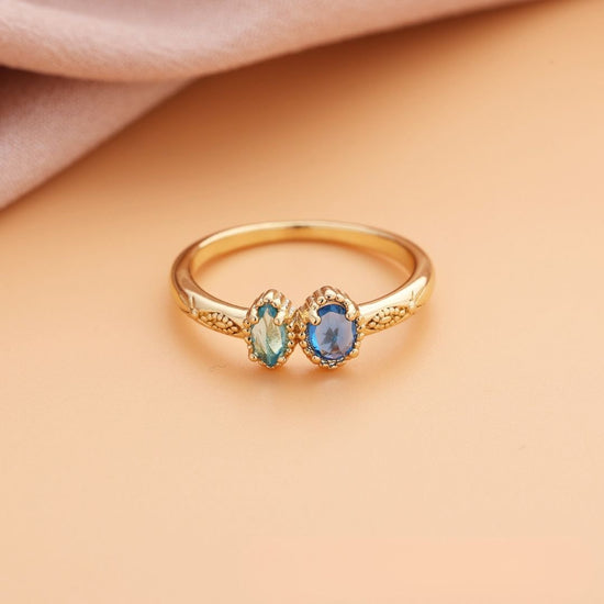 Anillo con piedras natales dorado con dos gemas ovales, una azul claro y otra azul intenso, juntas. Exhibido sobre fondo melocotón y tela rosa suave. Ideal como regalo de Día de las Madres.