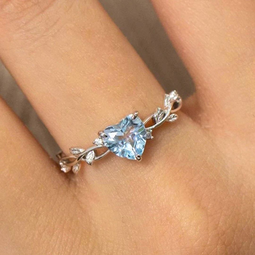 Primer plano de una mano que lleva el Anillo corazón del bosque, con una gema azul en forma de corazón en el centro y delicados detalles en forma de hoja en su correa ajustable.