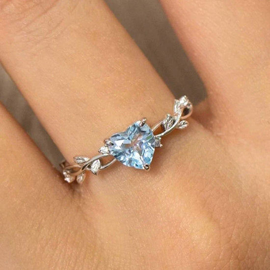 Primer plano de una mano que lleva el Anillo corazón del bosque, con una gema azul en forma de corazón en el centro y delicados detalles en forma de hoja en su correa ajustable.