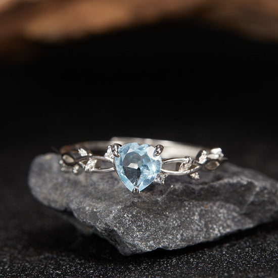 El Anillo corazón del bosque, un anillo plateado con una gema azul claro en forma de corazón y pequeñas piedras transparentes engastadas en una delicada banda de inspiración botánica, se exhibe sobre una roca gris oscura sobre un fondo difuminado.