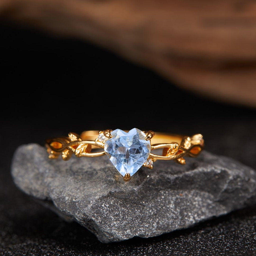 El Anillo corazón del bosque, con su tono dorado y su gema en forma de corazón adornada con intrincados diseños de hojas, descansa sobre una piedra oscura, capturando la esencia de la elegancia botánica.