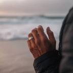 Primer plano de una mano que lleva el Anillo de Magnoterapia - Alivio para la menopausia, con las olas del mar de fondo. La persona, vestida con una chaqueta oscura, disfruta de la playa al atardecer o al amanecer.