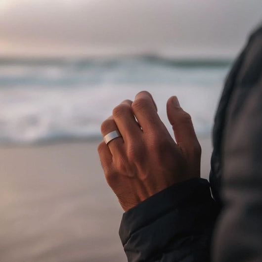 Primer plano de una mano que lleva el Anillo de Magnoterapia - Alivio para la menopausia, con las olas del mar de fondo. La persona, vestida con una chaqueta oscura, disfruta de la playa al atardecer o al amanecer.
