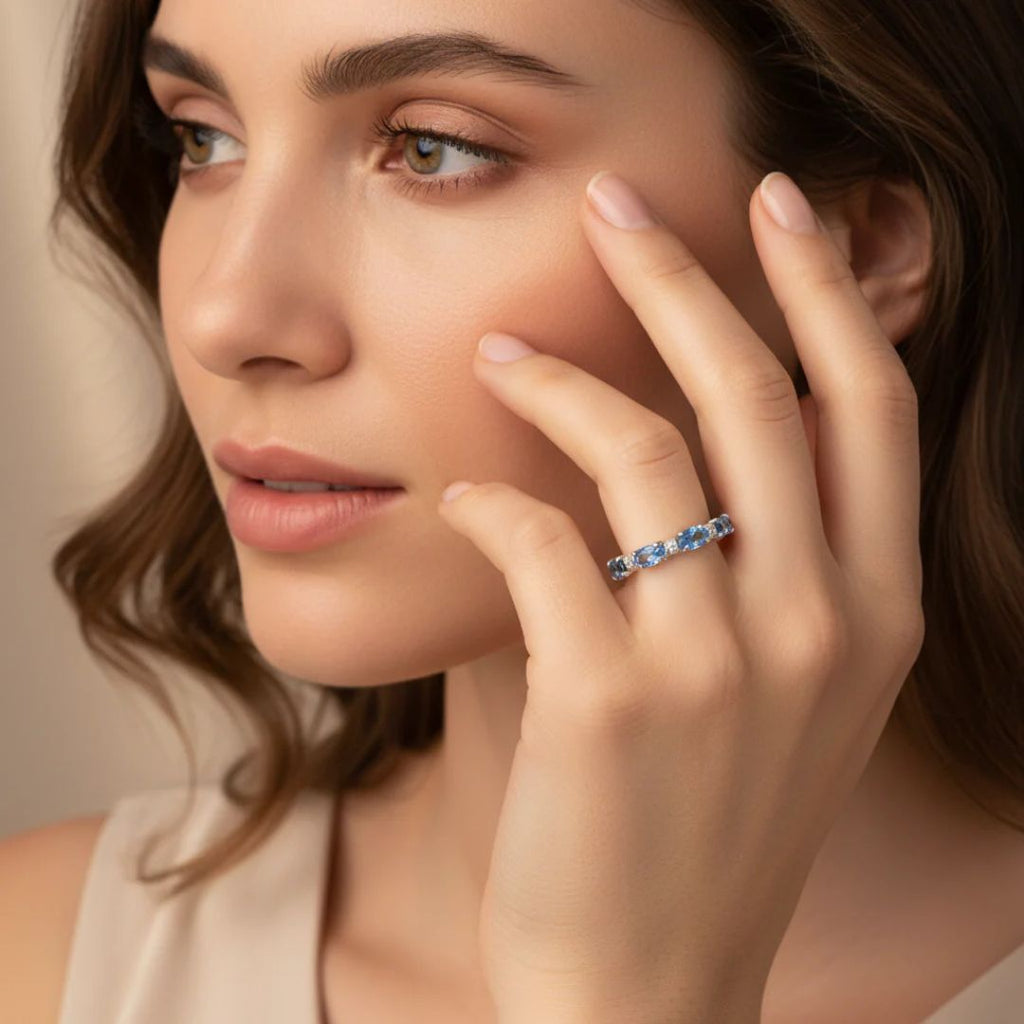 Una mujer de piel clara y pelo castaño se toca suavemente la cara, mostrando el Anillo Energía Zodiacal con piedra de nacimiento -un anillo de plata engastado con una piedra preciosa azul- en el dedo. Parece tranquila y lleva un top beige sin mangas.
