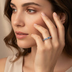 Una mujer de piel clara y pelo castaño se toca suavemente la cara, mostrando el Anillo Energía Zodiacal con piedra de nacimiento -un anillo de plata engastado con una piedra preciosa azul- en el dedo. Parece tranquila y lleva un top beige sin mangas.