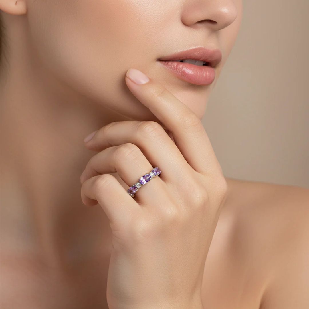 Una mujer de piel suave se toca la barbilla mostrando el Anillo Energía Zodiacal con piedra de nacimiento, un anillo de plata con una gema morada. El fondo beige neutro resalta el encanto de esta joyería espiritual.