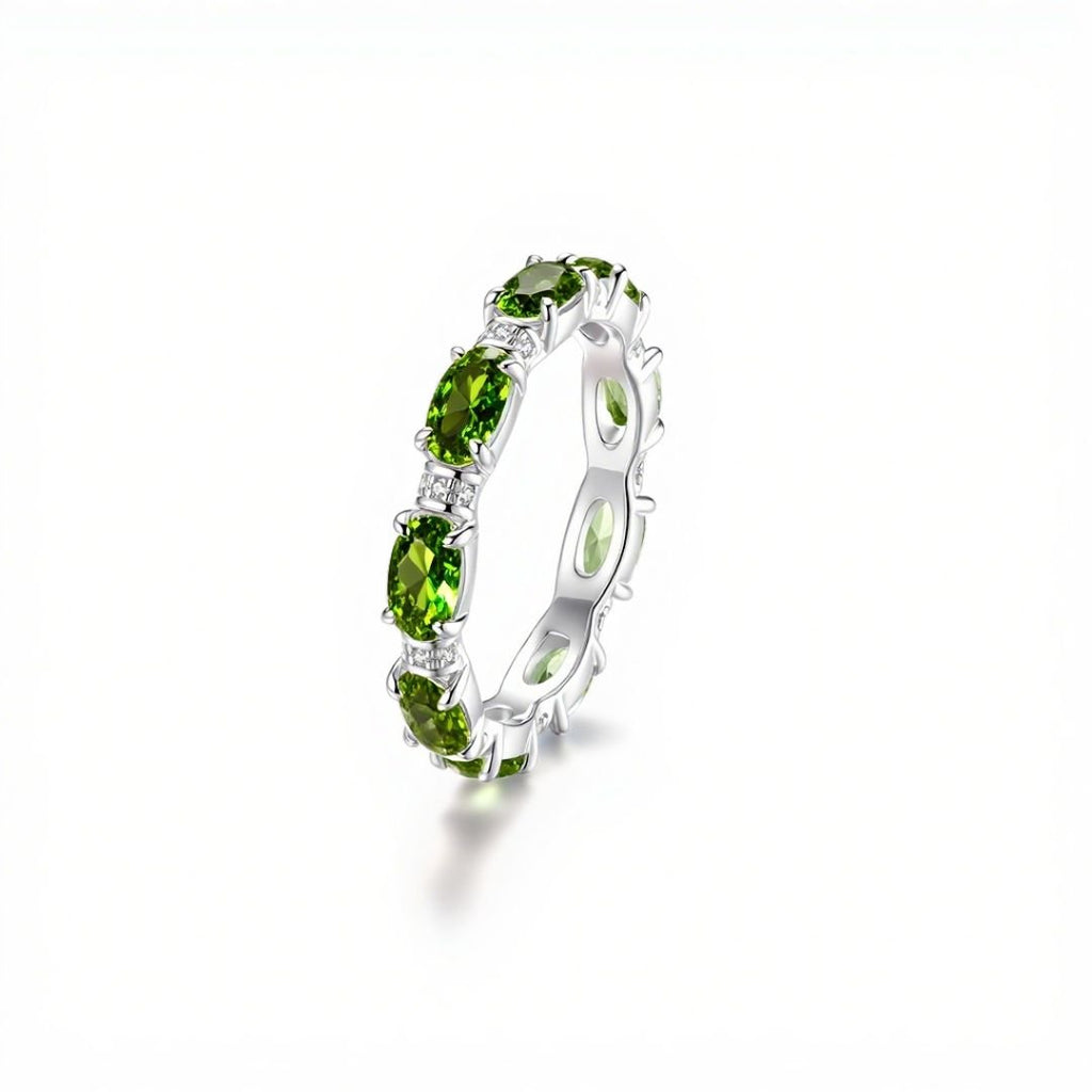 El Anillo Energía Zodiacal con piedra de nacimiento es una banda plateada con gemas verdes ovaladas engastadas en grifas, conocidas por sus propiedades energizantes en la joyería espiritual, mostrada sobre fondo blanco.