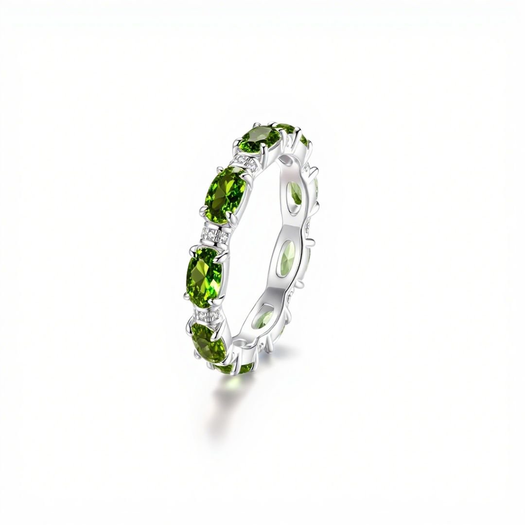 El Anillo Energía Zodiacal con piedra de nacimiento es una banda plateada con gemas verdes ovaladas engastadas en grifas, conocidas por sus propiedades energizantes en la joyería espiritual, mostrada sobre fondo blanco.