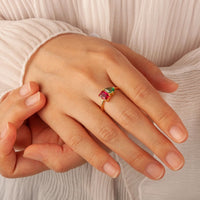 Primer plano de una mano que lleva el Anillo Eterno con piedras de nacimiento (Oferta 2x1), adornado con coloridas piedras de nacimiento, sobre un ligero tejido plisado. Otra mano toca suavemente el dedo anular, un regalo perfecto para parejas.