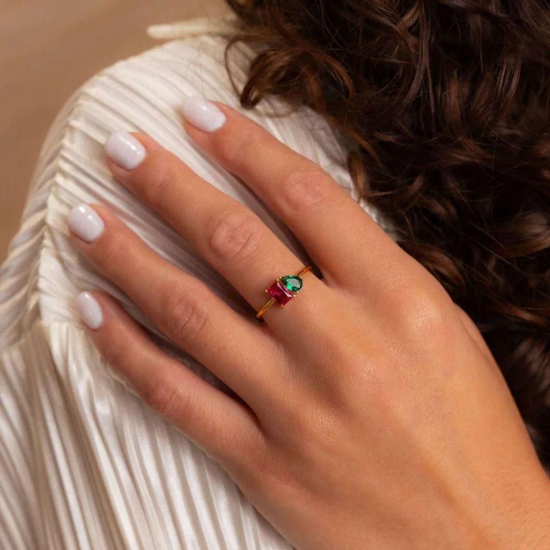 Una mano con las uñas cuidadas y pintadas de blanco descansa sobre una tela blanca plisada. La persona lleva un Anillo Eterno con piedras de nacimiento (Oferta 2x1), ideal como regalo para parejas. Al fondo se ve el pelo castaño rizado.