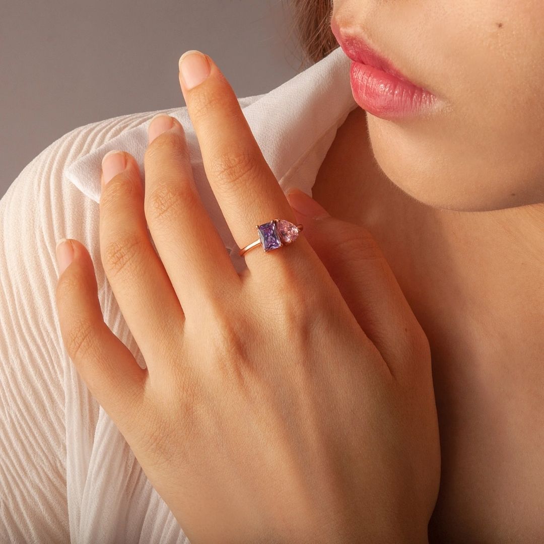 Una mujer con blusa blanca se toca la cara, mostrando el Anillo Eterno con piedras de nacimiento (Oferta 2x1), con una gran piedra preciosa púrpura y una piedra rosa en forma de pera-un elegante regalo para parejas o para celebrar los nacimientos.